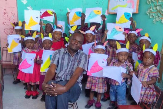 Students and their teacher in a Cité Soleil classroom holding up their paper artwork