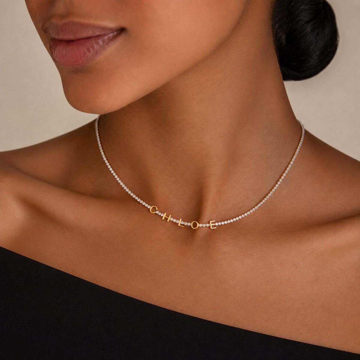 Name tennis necklace spelling CHLOE worn at the collarbone over a black off-shoulder top