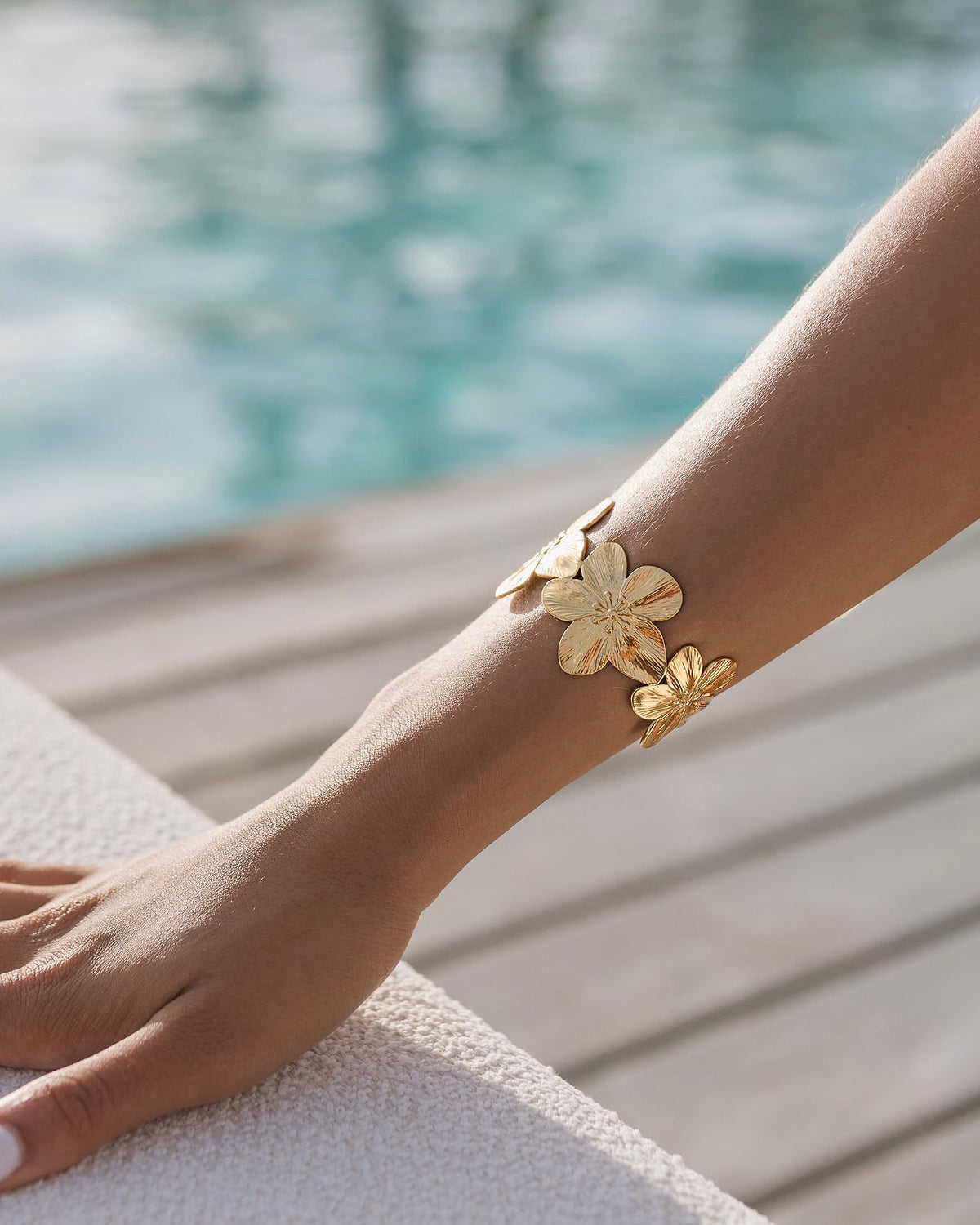 Gold flower cuff bracelet worn on the wrist beside a pool